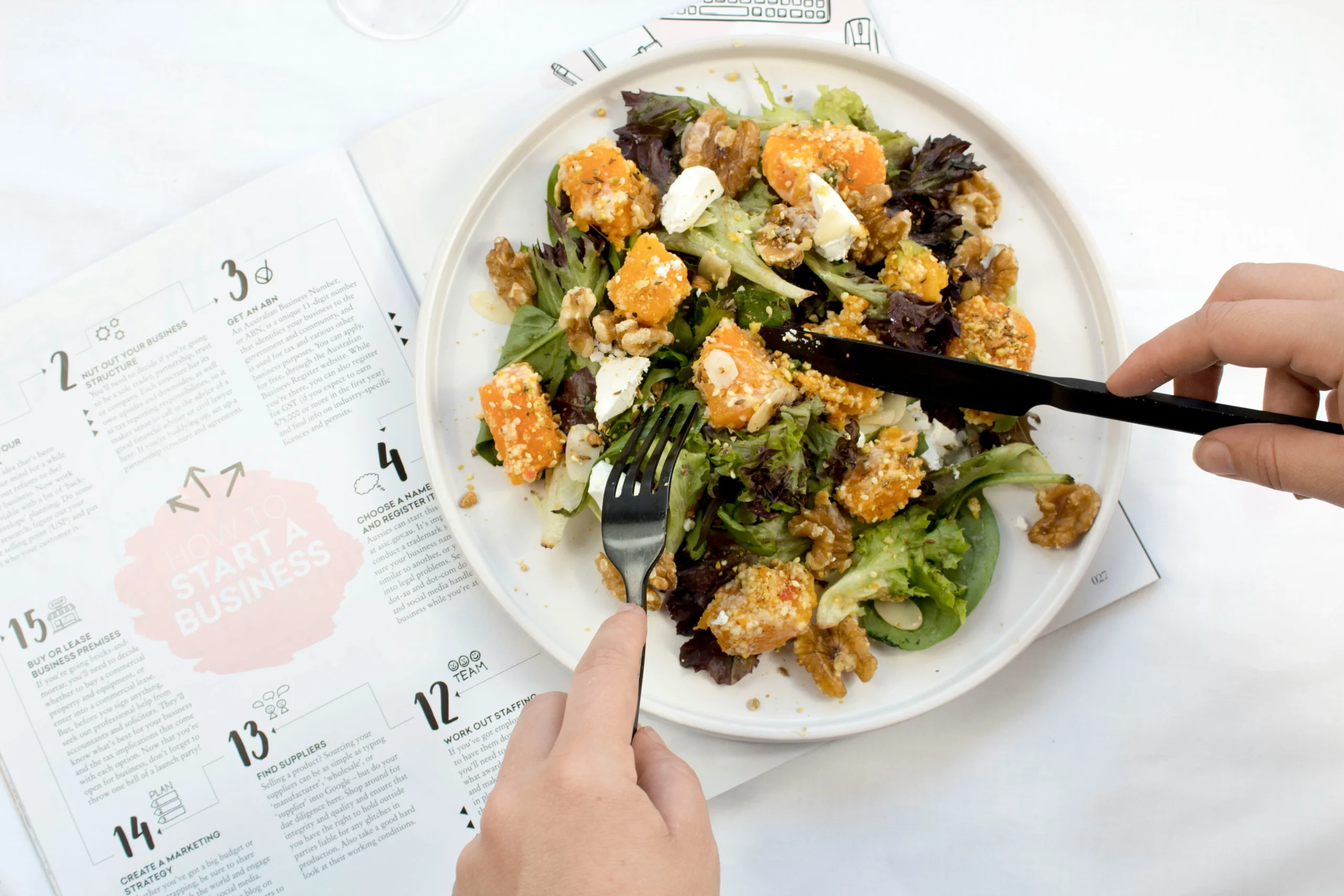 A person is using a black knife and fork to eat a colorful salad on a white plate. The salad consists of mixed greens, chunks of roasted butternut squash, walnuts, and crumbled cheese. The plate is placed on top of an open book or magazine that discusses conversion rate optimisation and business startup steps, visible with numbered sections and text. The background is light and airy, creating a fresh and productive atmosphere, blending healthy eating with business planning and conversion rate optimisation strategies.