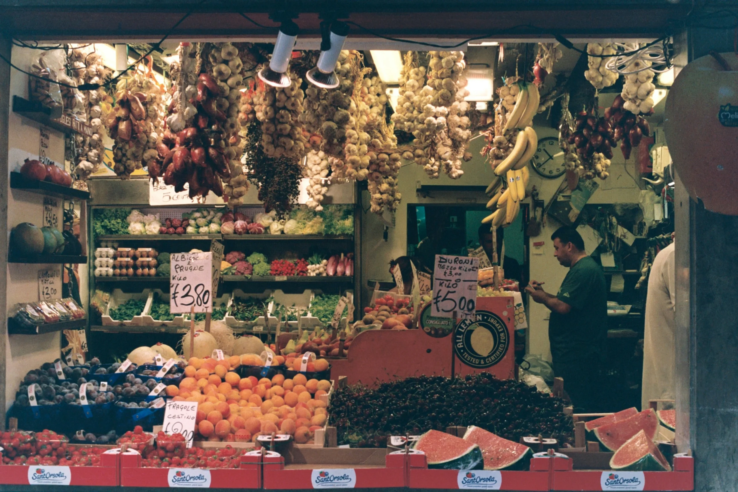 A bustling market stall displaying a variety of fresh fruits and vegetables, including bananas, watermelons, peaches, and garlic. The vendor is seen taking notes, possibly conducting market research on customer preferences